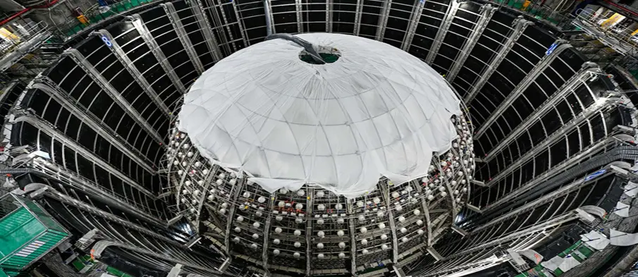 The JUNO main detector 750m underground in a dedicated laboratory. The photo shows the (still empty) water pool of the detector with the central scaffolding. Inside this globe, the acrylic sphere with 34.5m diameter is placed, filled with the liquid scintillator. The white cover just protects sensitive components during mounting.